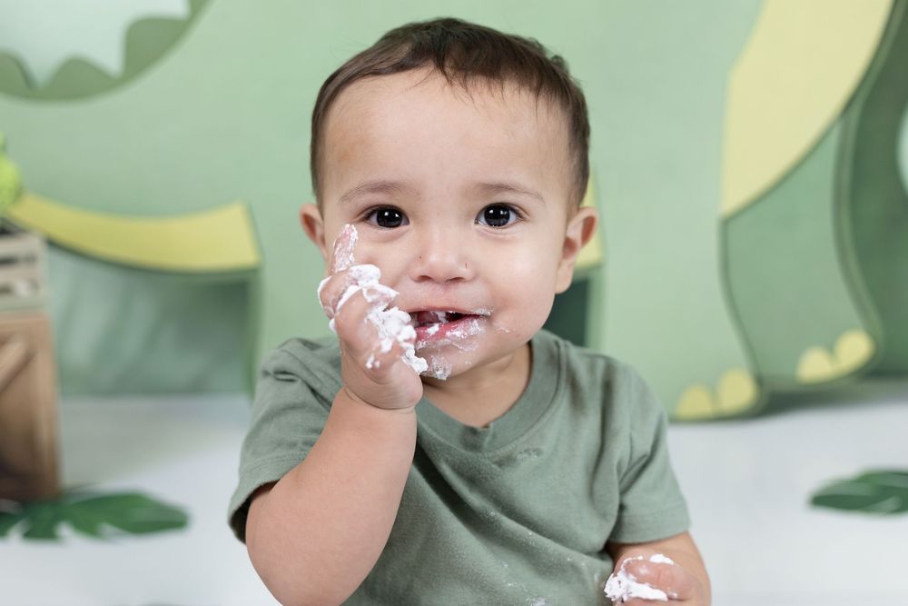 Baby boy in green onesie enjoys smashing birthday cake during first birthday cake smash photo session.