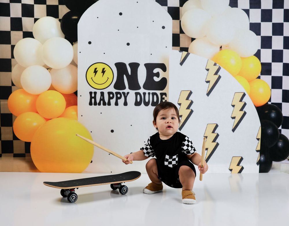 Baby boy sits with skateboard at ONE HAPPY DUDE first birthday party with balloon arch backdrop.
