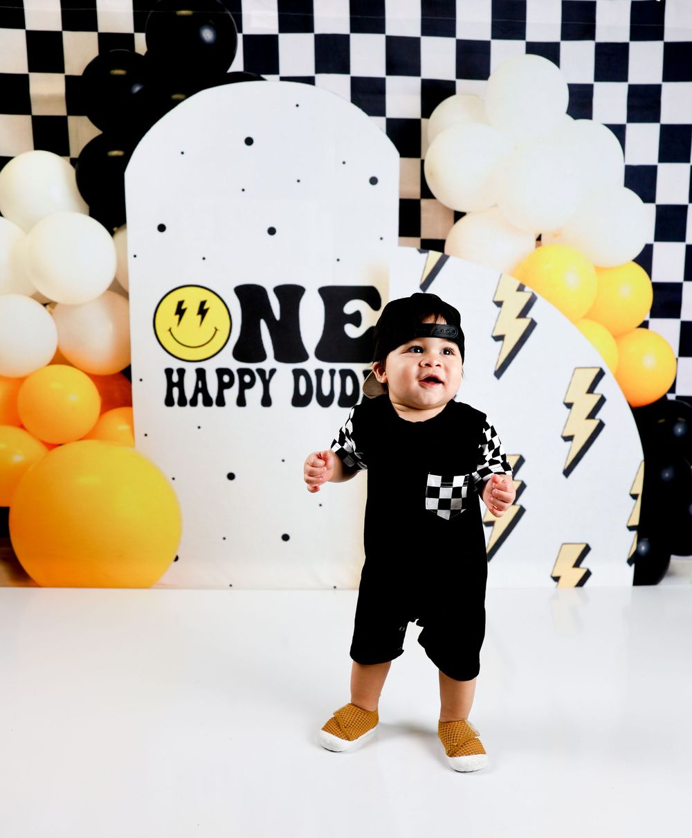 Toddler boy in checkered outfit stands before ONE HAPPY DUDE birthday backdrop with black, white, and yellow balloons.