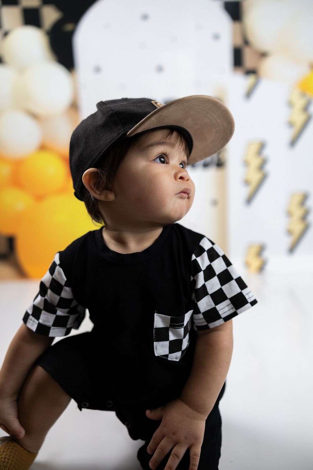Adorable toddler boy in black checkered shirt and cap sitting against festive yellow balloon backdrop.