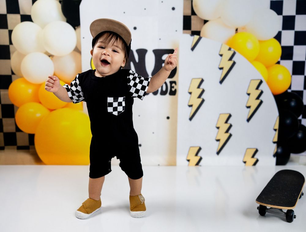 Toddler boy in checkered romper celebrating first birthday with balloons and lightning bolt decorations.