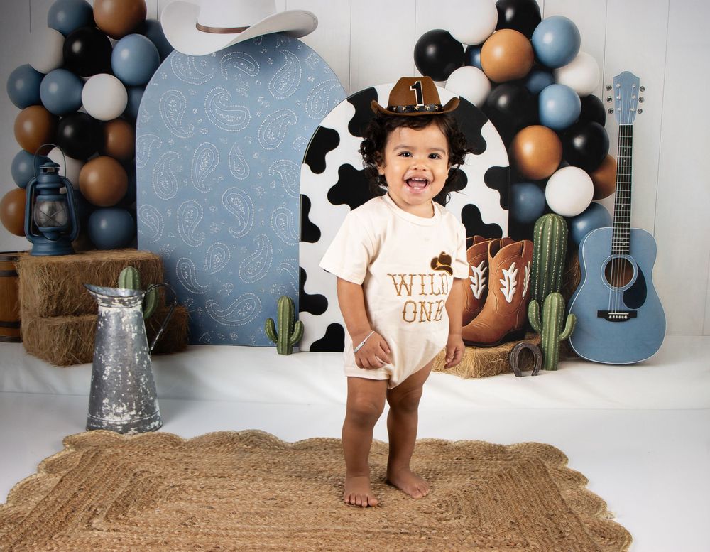 Baby in cowboy hat wearing Wild One onesie at first birthday photoshoot with western balloon backdrop.