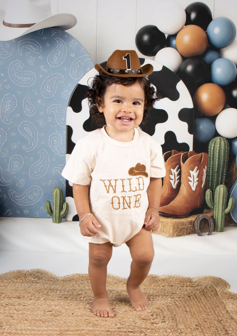 Smiling baby in cowboy hat and Wild One onesie celebrates first birthday with western themed balloon backdrop.