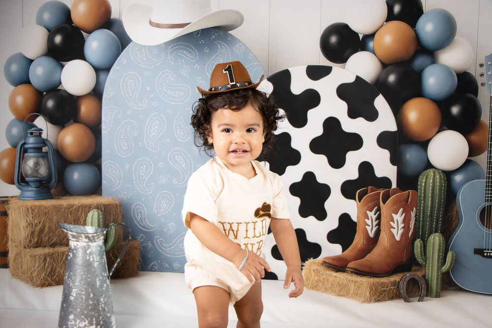 Toddler in cowboy hat and white onesie poses at a Western-themed first birthday setup with balloons and boots.