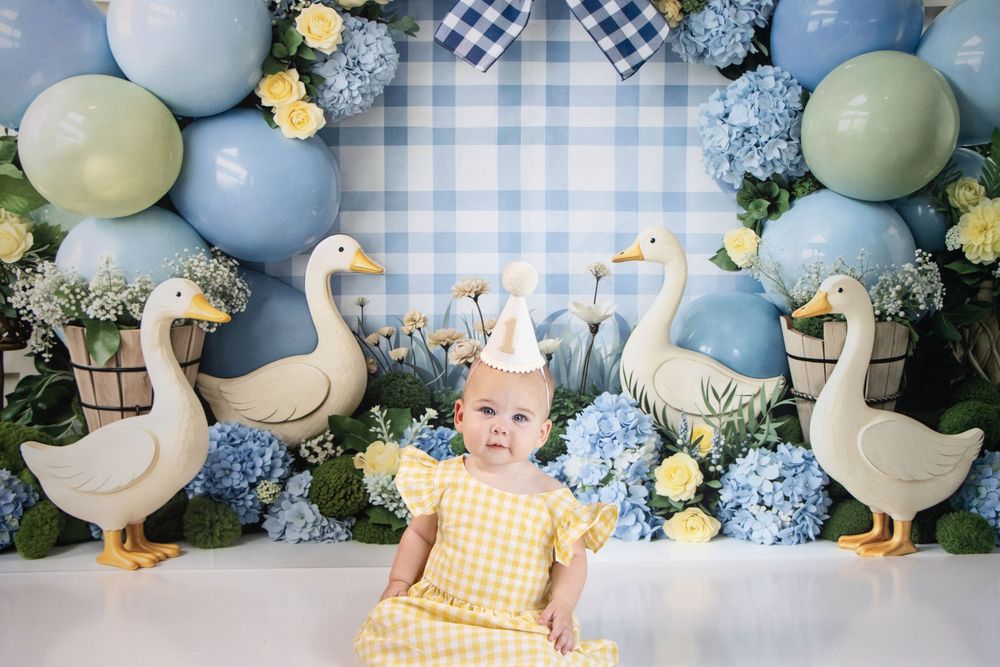 Baby in yellow dress sits before blue hydrangeas, balloons, and goose decorations for a first birthday cake smash.