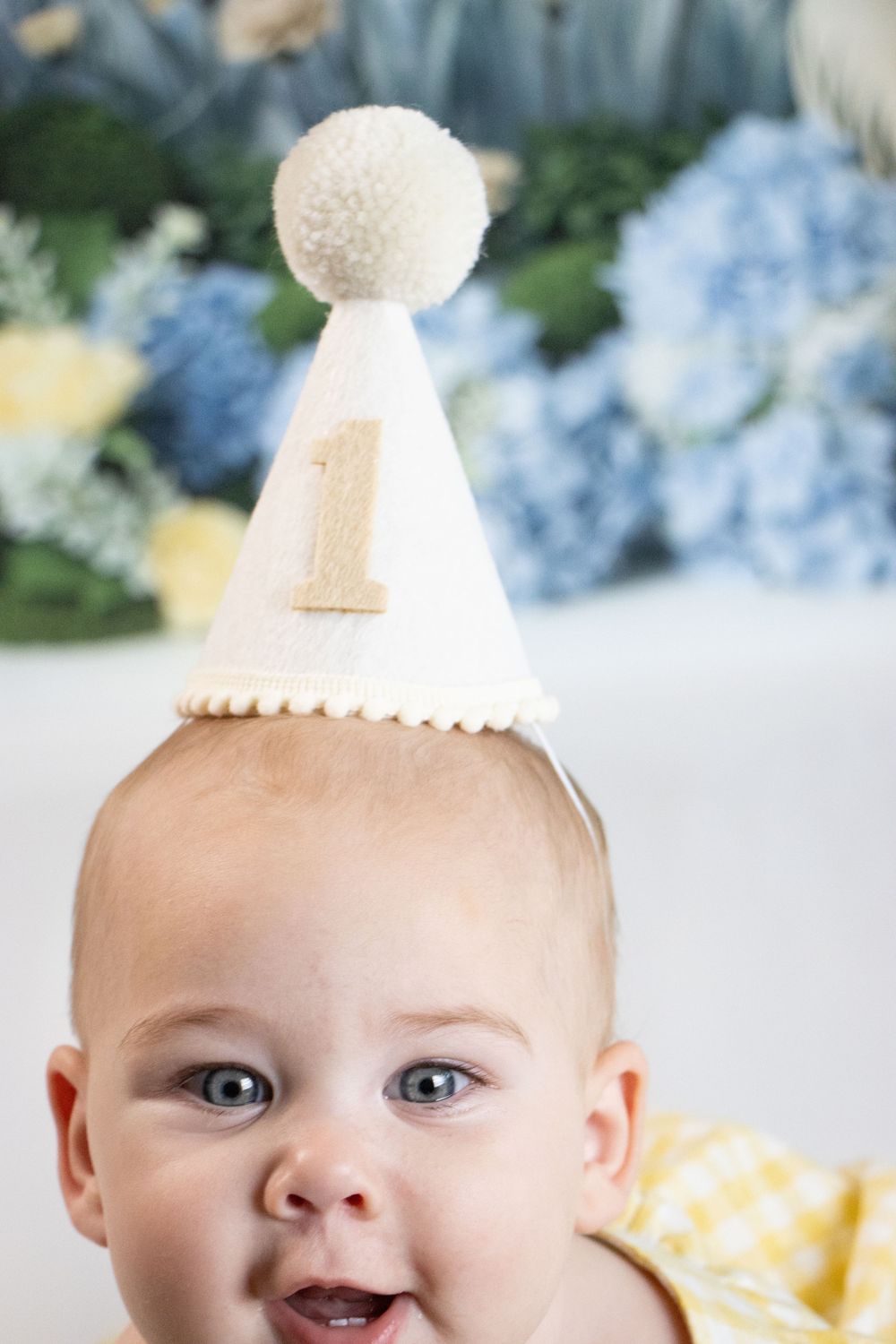 Smiling baby wearing a white felt first birthday party hat with pom pom and number 1, with floral backdrop.