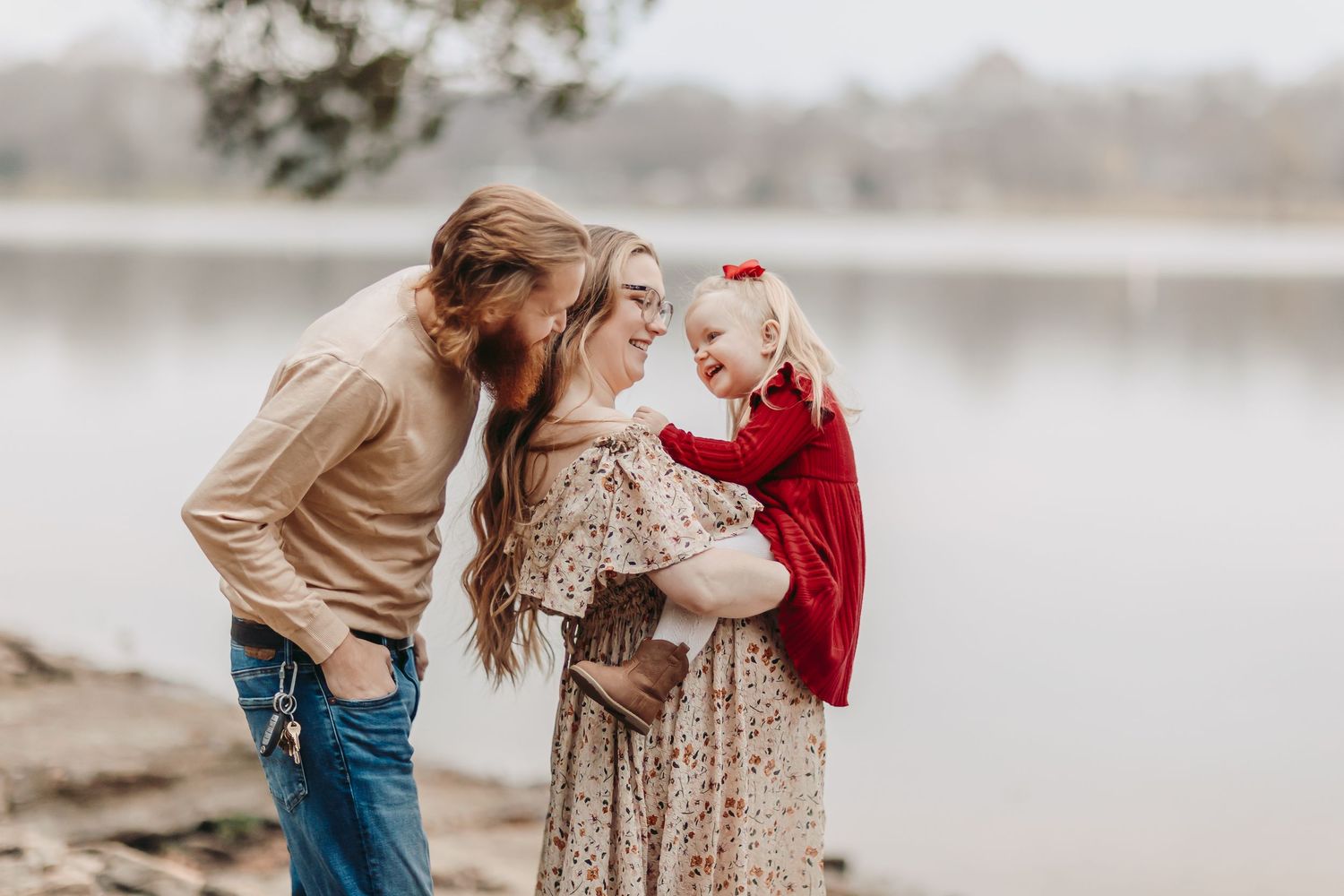 A young family with a toddler girl in red shares a joyful moment by a serene lake during a fall photo session.
