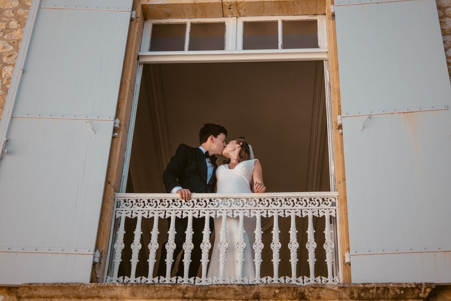 Beth and Matt kissing on the château balcony between pale blue shutters.