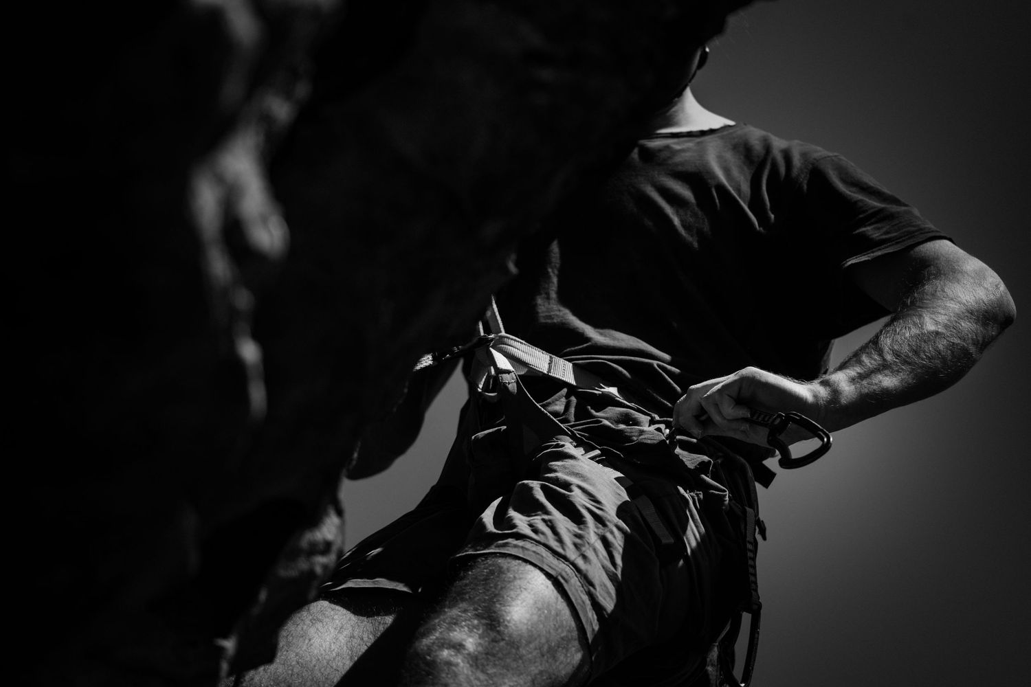 Black and white photo of a rock climber adjusting climbing gear on a cliff face.