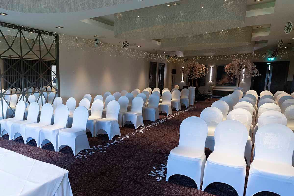 Elegant wedding ceremony venue with white chair covers arranged in rows on patterned carpet with floral decorations.