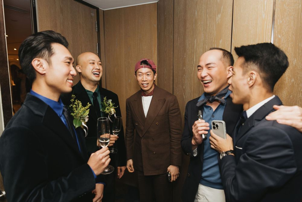 Five men in formal attire socializing and laughing at an indoor event, some holding drinks and phones.