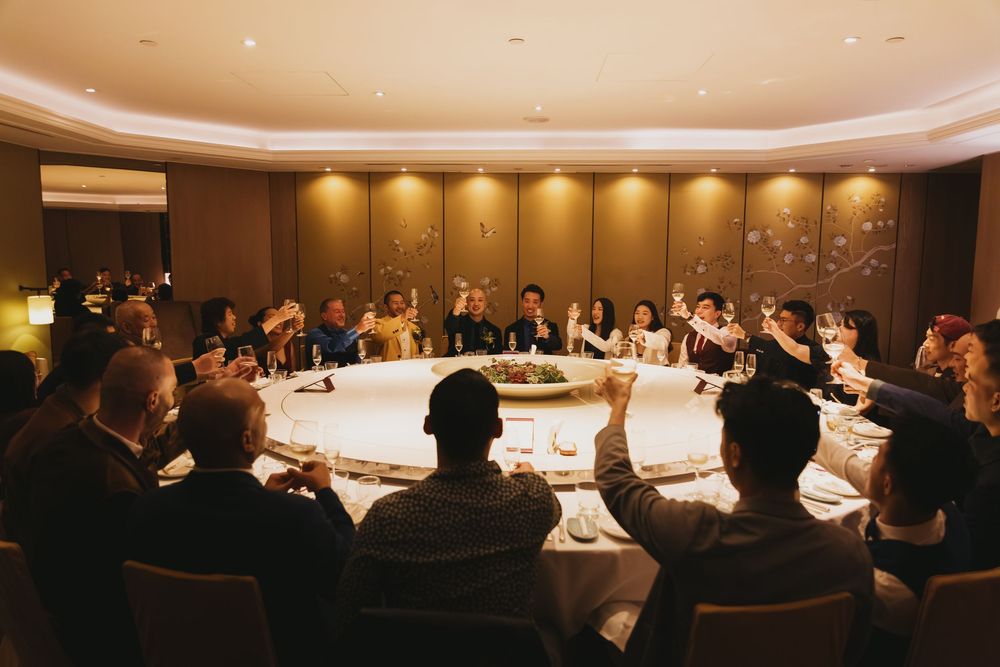 Elegant group dinner toast at a round table in a warmly lit private dining room with guests raising glasses.