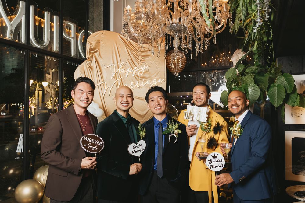 Five men in formal attire smile and hold photo booth props at an elegant venue with chandeliers and gold decor.