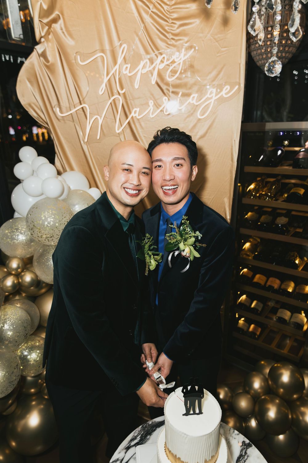 Two grooms smile while cutting their wedding cake in front of a Happy Marriage neon sign with gold balloons.