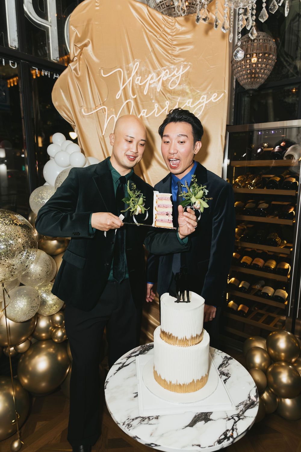 Two grooms in green velvet suits pose happily beside their wedding cake at a gold-themed celebration.