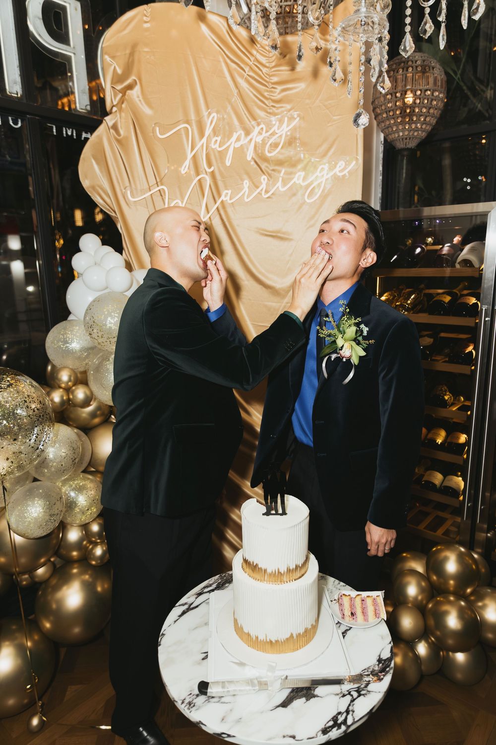 Two grooms feed each other wedding cake at a gold-themed reception with balloon decor and a Happy Marriage sign.