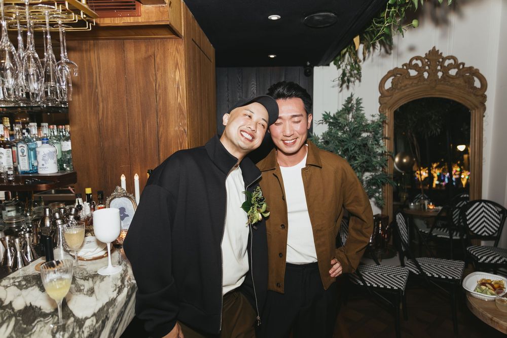 Two smiling men pose together at a stylish bar with cocktails, bottles, and elegant decor in the background.