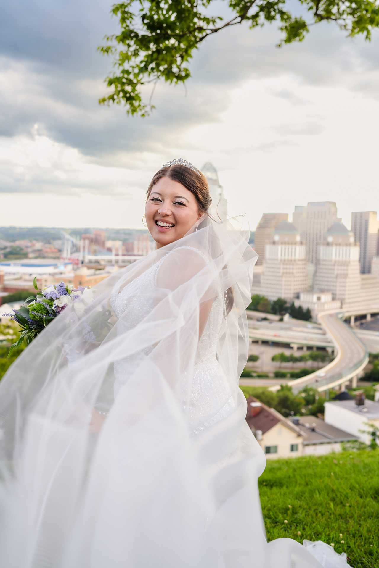 Smiling bride in white gown and flowing veil holds bouquet outdoors with city skyline backdrop on cloudy day at the view wedding venue