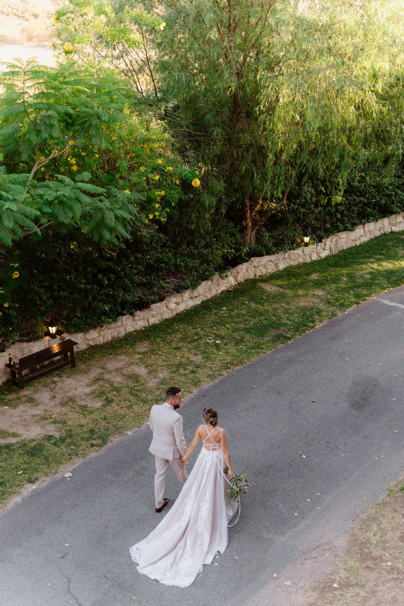 Bride and groom walk hand-in-hand down a quiet road, surrounded by greenery, captured from above in a peaceful moment.