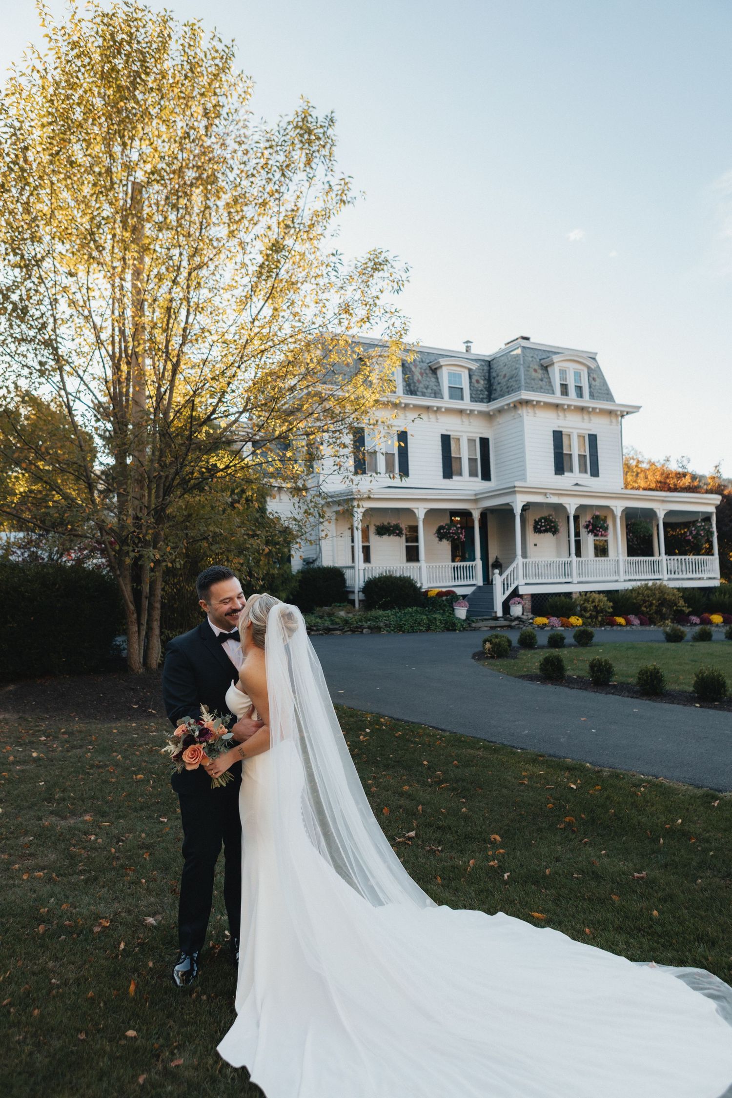 Bride and groom share a kiss on the lawn of a white Victorian estate during an autumn wedding.