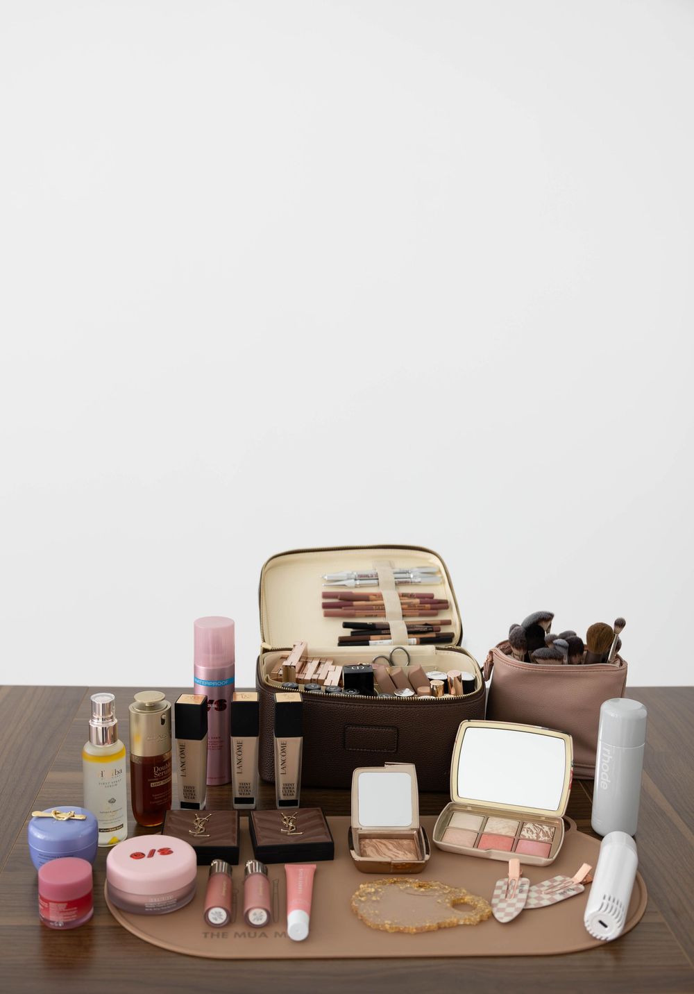 Brown makeup travel case open on a wooden tray surrounded by luxury beauty products, brushes, jewelry, and cosmetics.