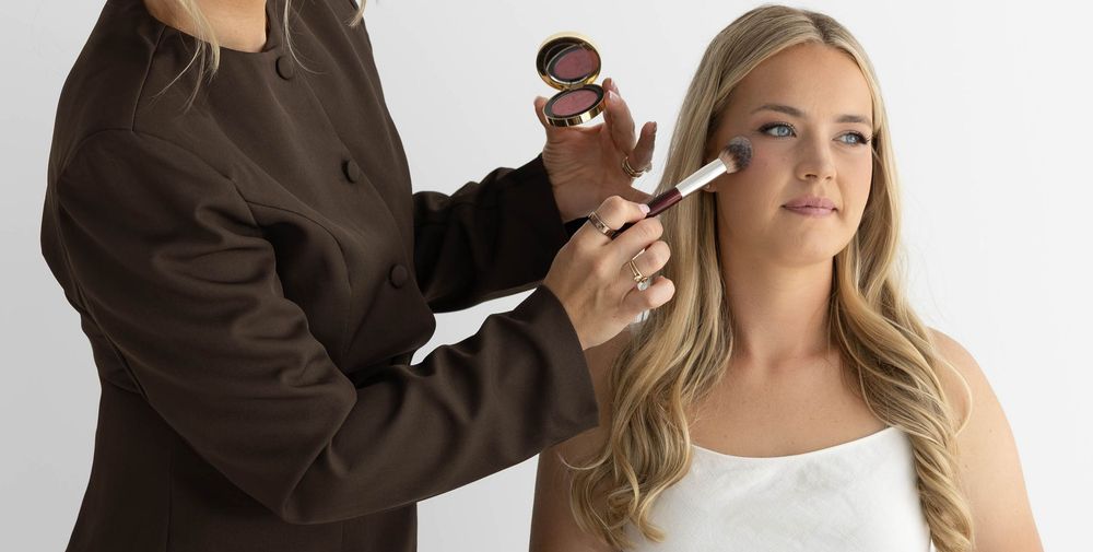 Makeup artist applying blush to a blonde woman's cheek using a brush and compact powder.