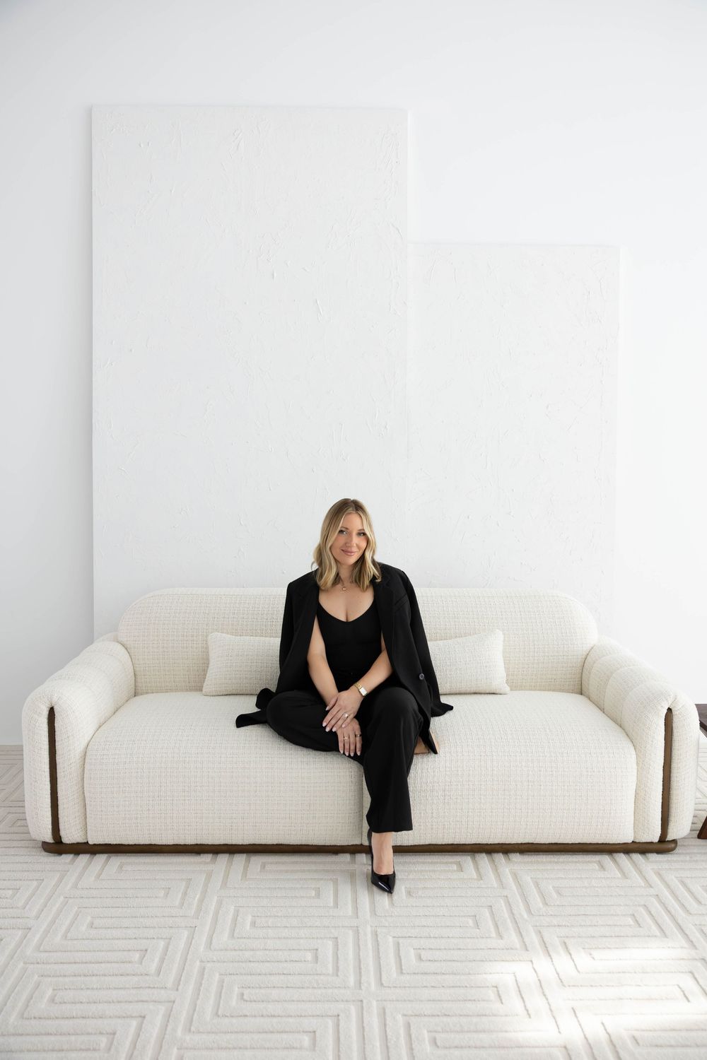 Woman in black outfit sitting on a white boucle sofa in a minimalist white room with geometric patterned rug.