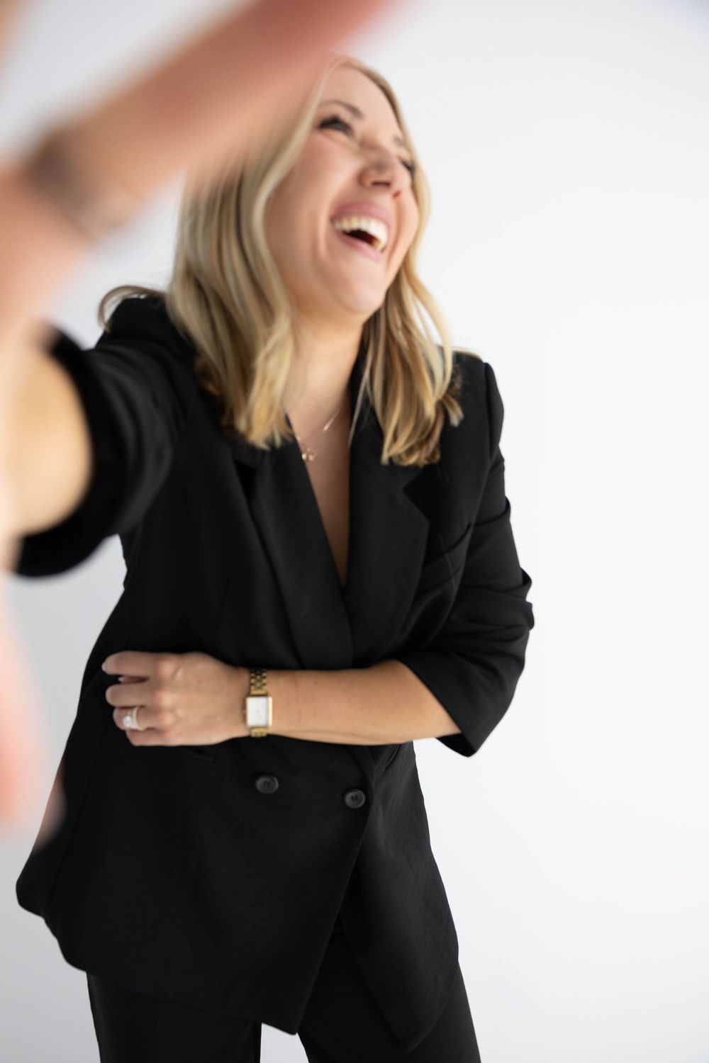 Laughing blonde woman in black blazer suit with gold watch, posing confidently against white background.