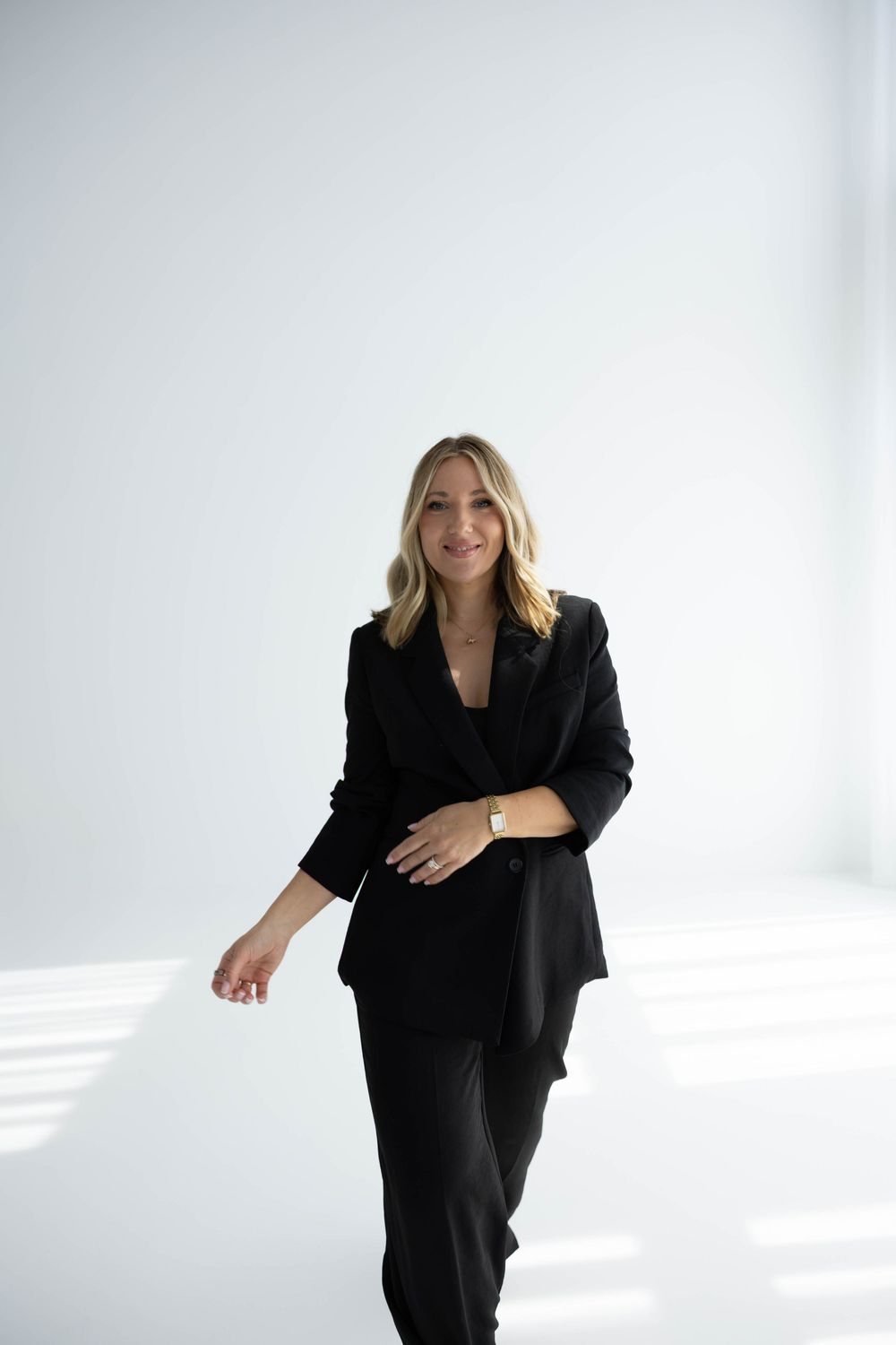 Confident blonde woman in black suit smiling and posing in bright white minimalist studio.