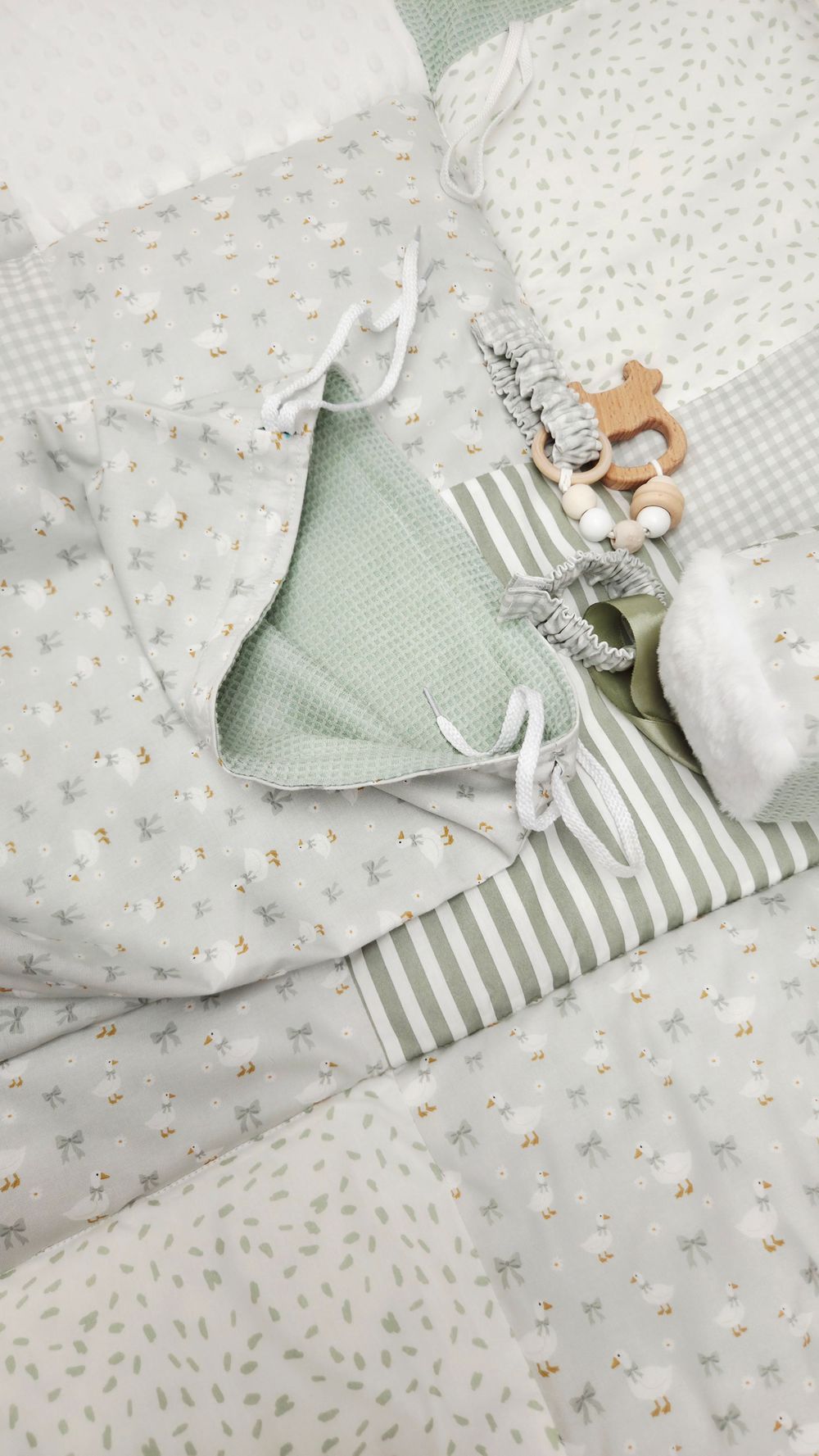 Soft sage green baby bedding set with floral and striped patterns, wooden teething toys, and white ribbon ties.