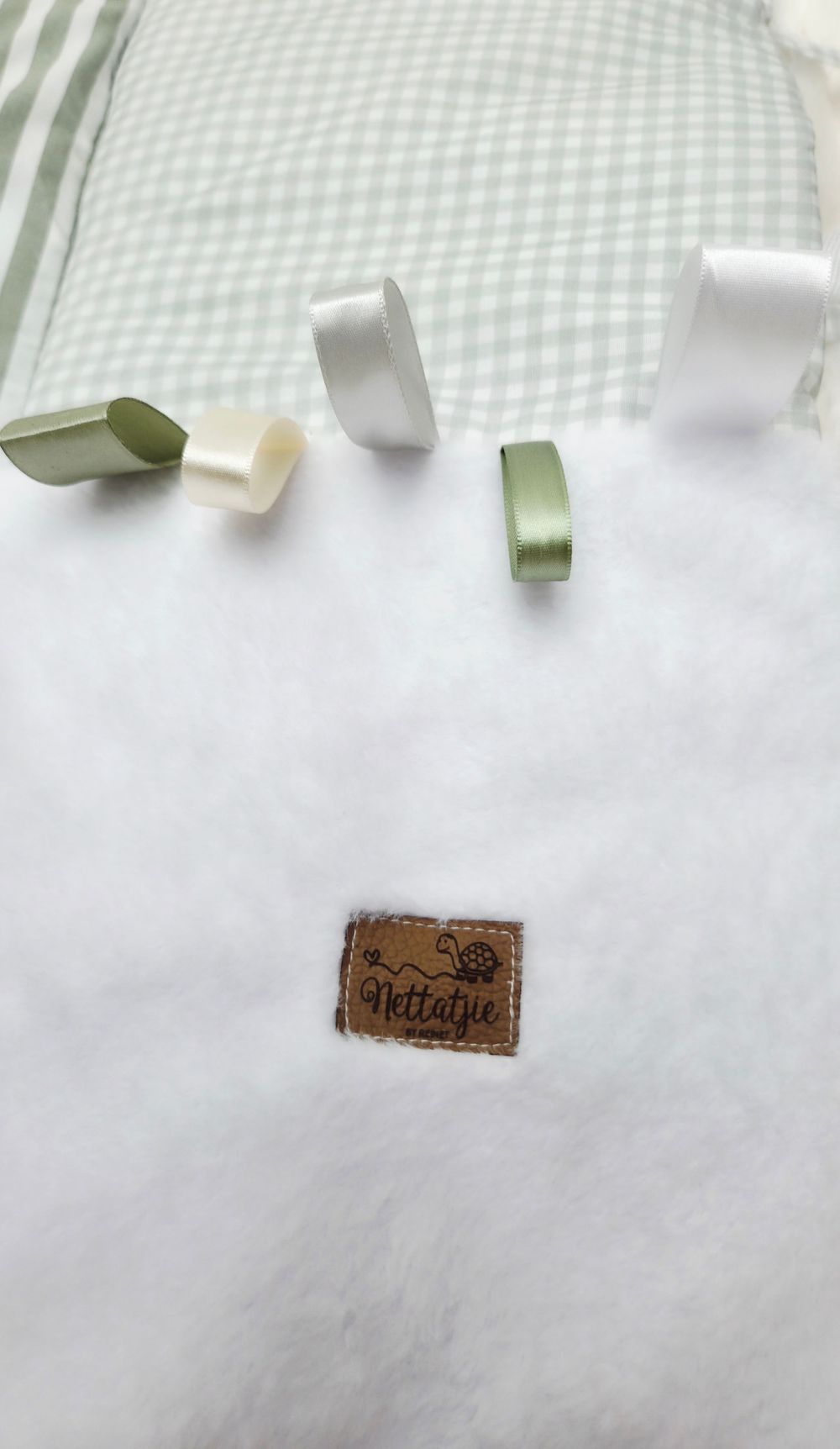 White fluffy baby blanket with green gingham trim, satin ribbons, and a brown Nettafie leather label.