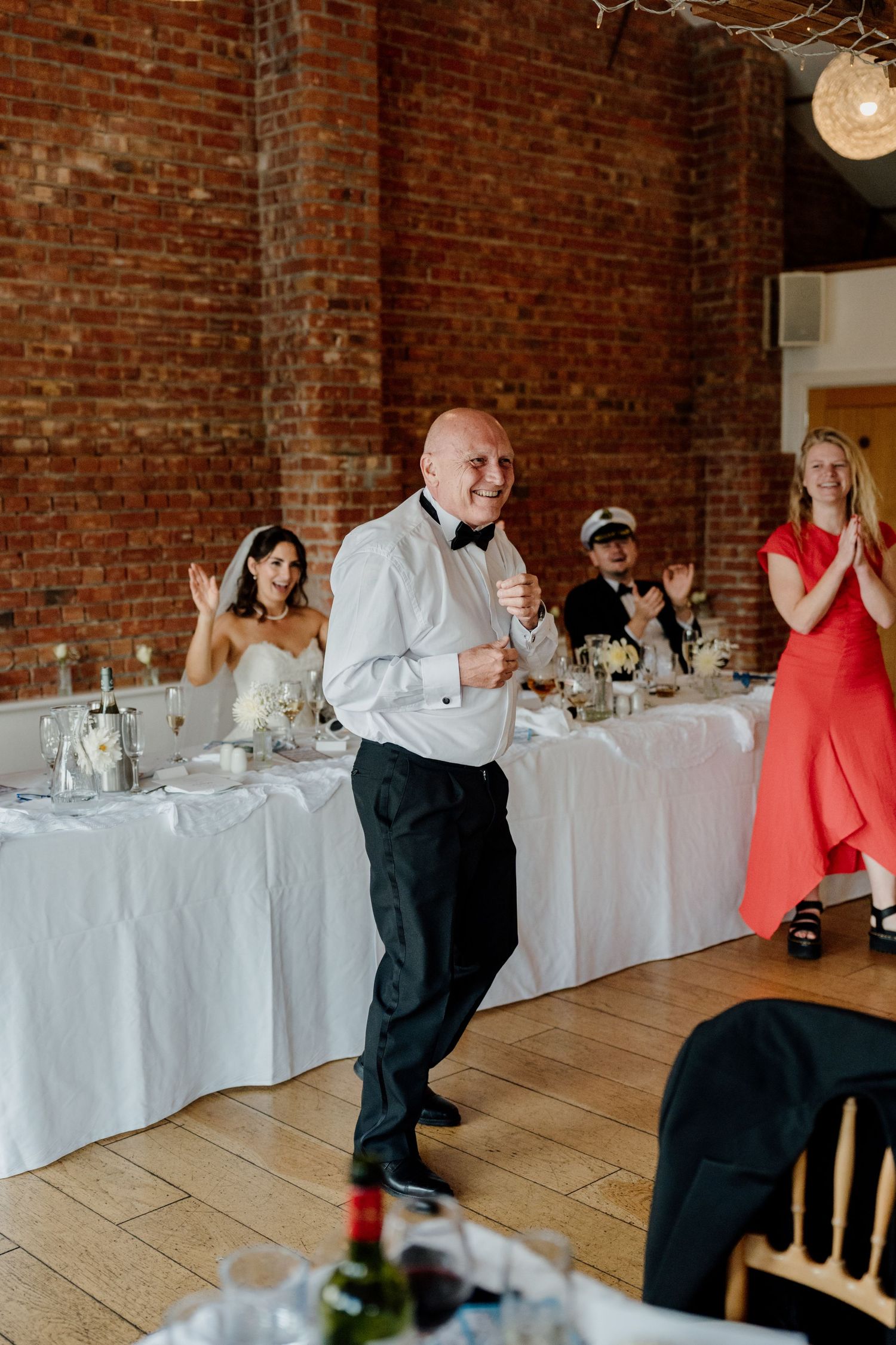 Bald man in tuxedo dancing joyfully at wedding reception in brick venue while guests applaud at decorated head table.