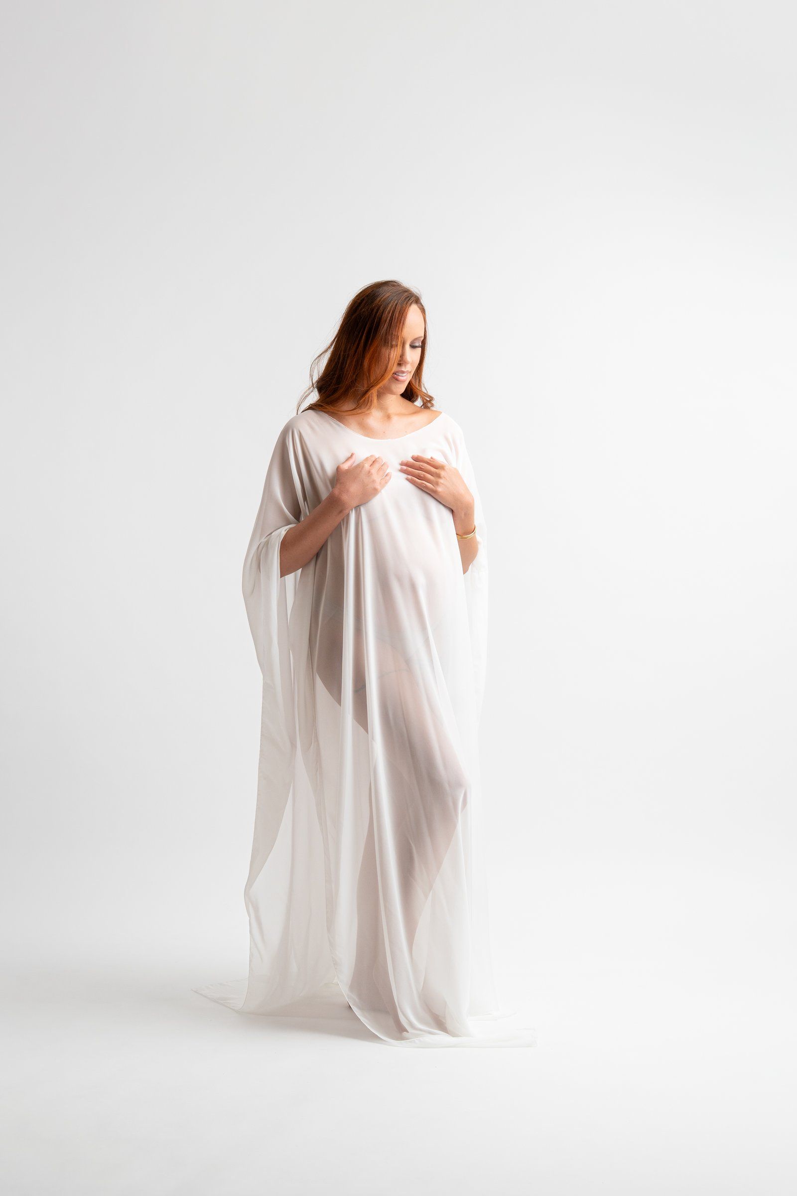 Pregnant woman in flowing white sheer fabric gown posing gracefully against a white background in a maternity photo.