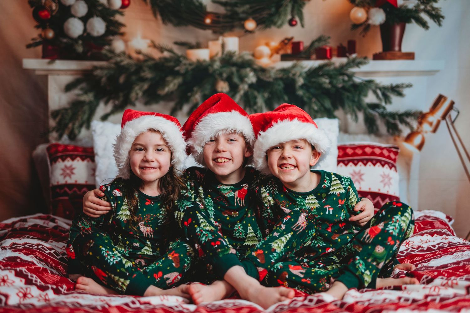 Three children in matching Christmas tree pajamas and Santa hats smile together on a bed near a decorated fireplace.