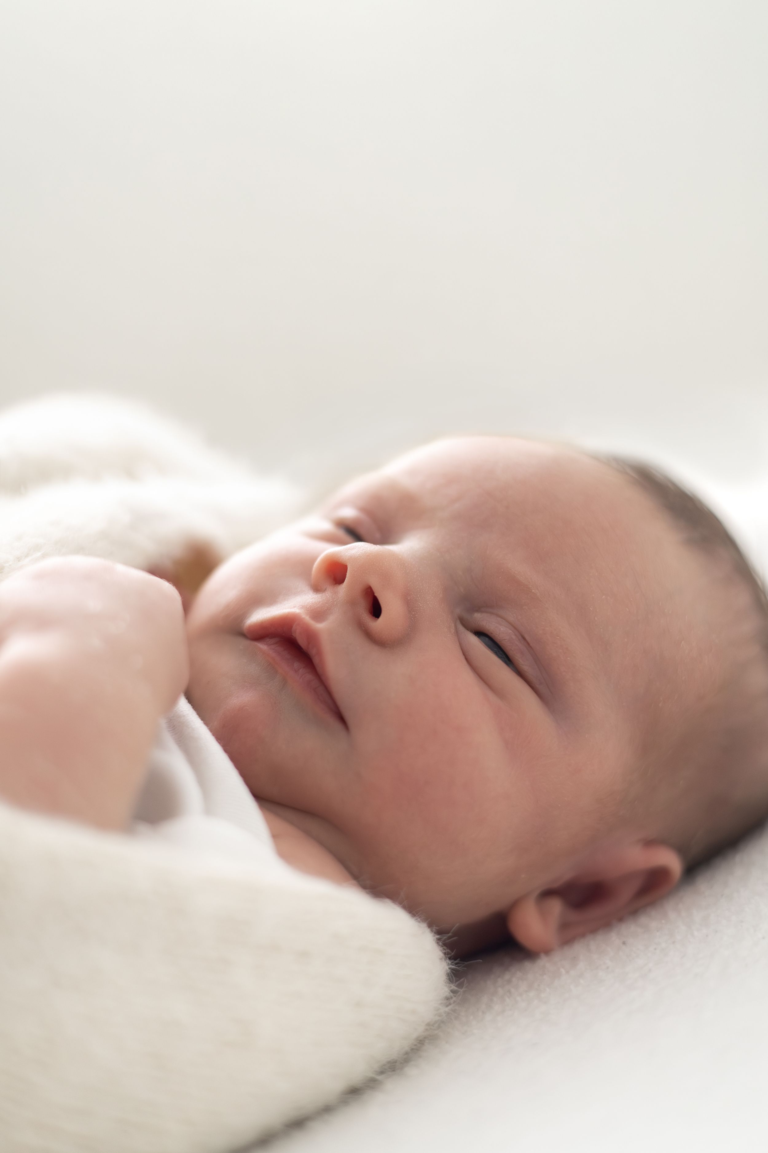 A peaceful newborn baby sleeping wrapped in a soft white blanket, with tiny hands near face.