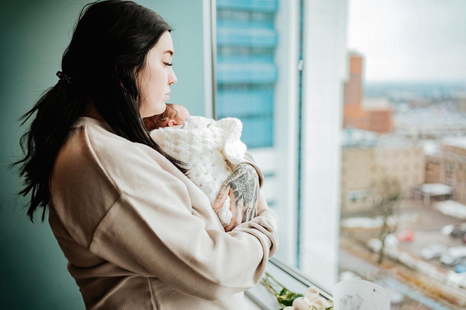 A young woman in a beige robe cradles her newborn baby by a hospital window overlooking a city.