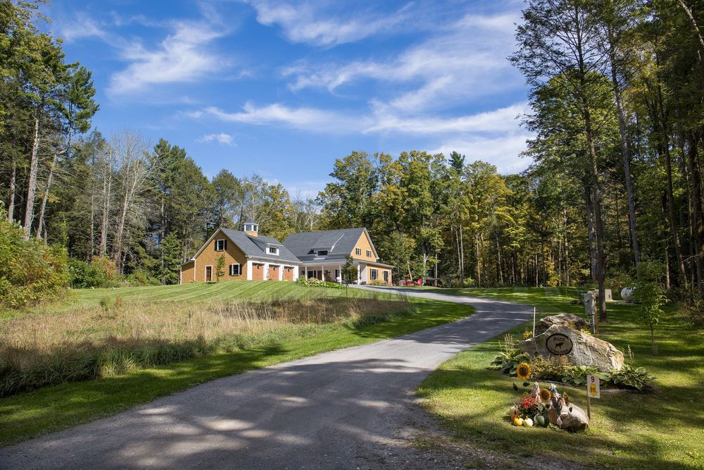 Rustic New England home with gravel driveway, autumn trees, and decorative pumpkin display at the entrance.