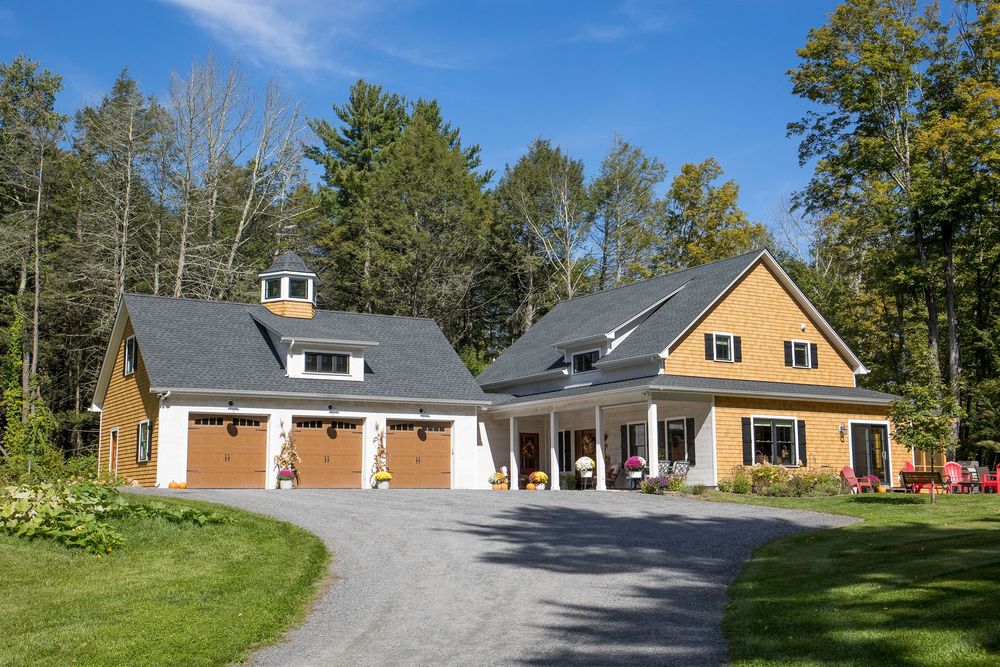 Elegant craftsman farmhouse with three-car garage, cedar siding, and lush green lawn surrounded by autumn trees.
