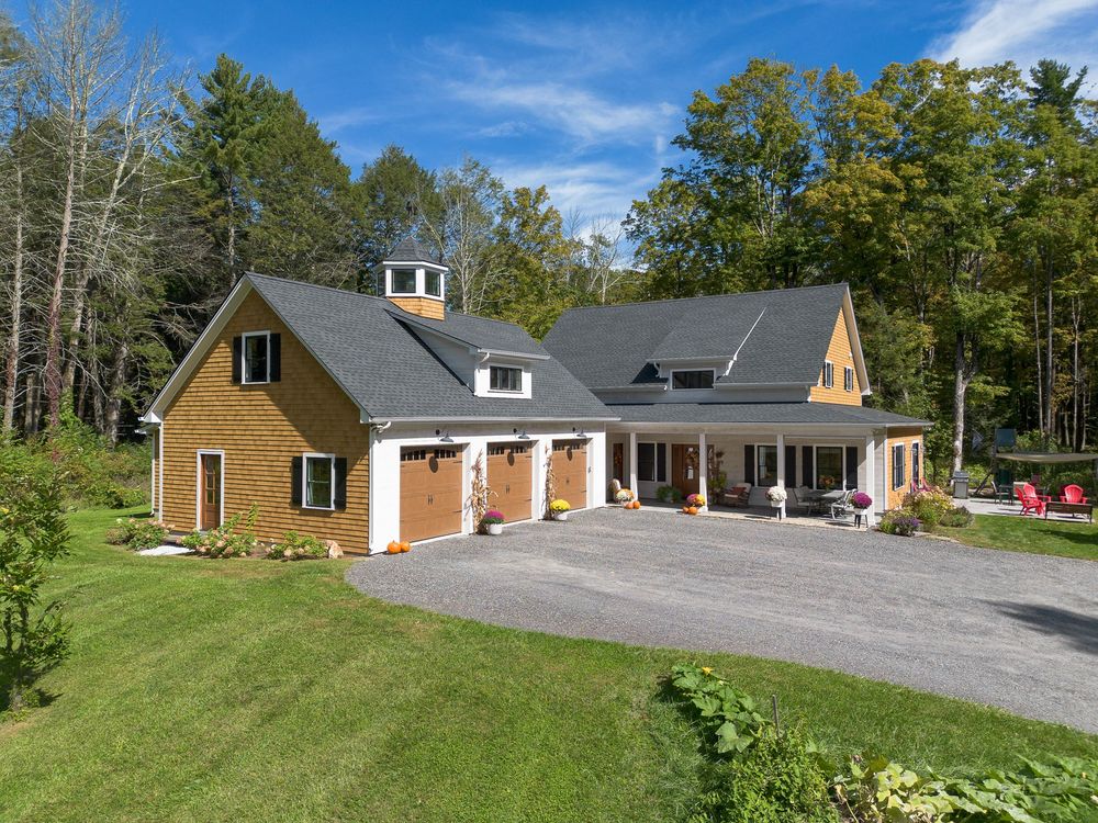 Stunning cedar-sided craftsman home with three-car garage and spacious covered porch amid lush New England forest.