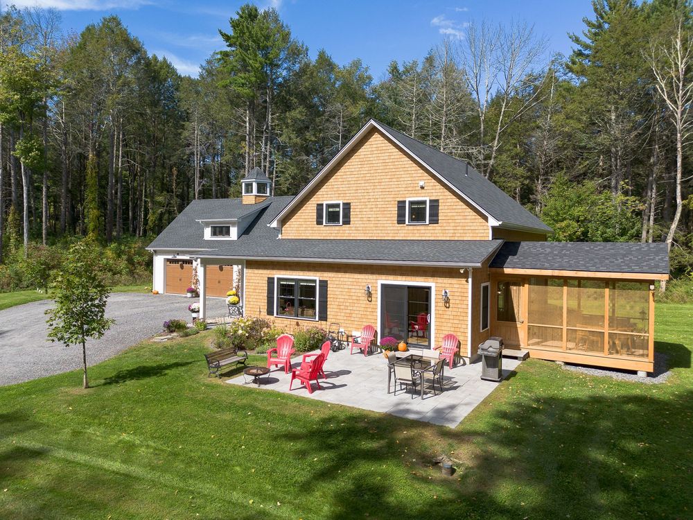 Aerial view of a cedar-shingled home with a screened porch, patio seating, and red Adirondack chairs surrounded by lush forest.