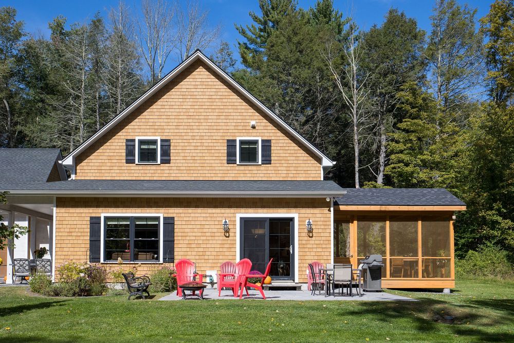 Charming cedar-shingled house with a screened porch, red Adirondack chairs, and a well-manicured lawn backed by autumn trees.