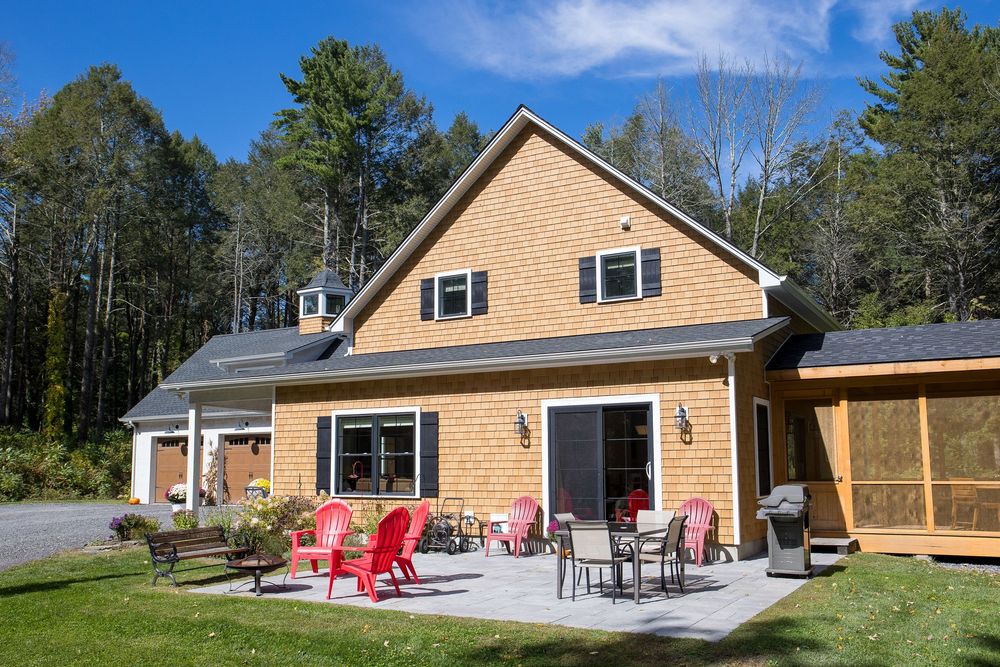 Cedar-sided New England home with a spacious patio featuring red Adirondack chairs and outdoor dining set under blue skies.