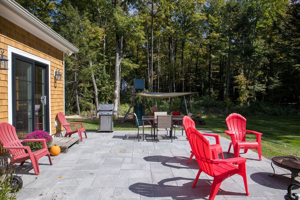 Spacious stone patio with bright red Adirondack chairs, outdoor dining set, and a grill beside a cedar-sided home.