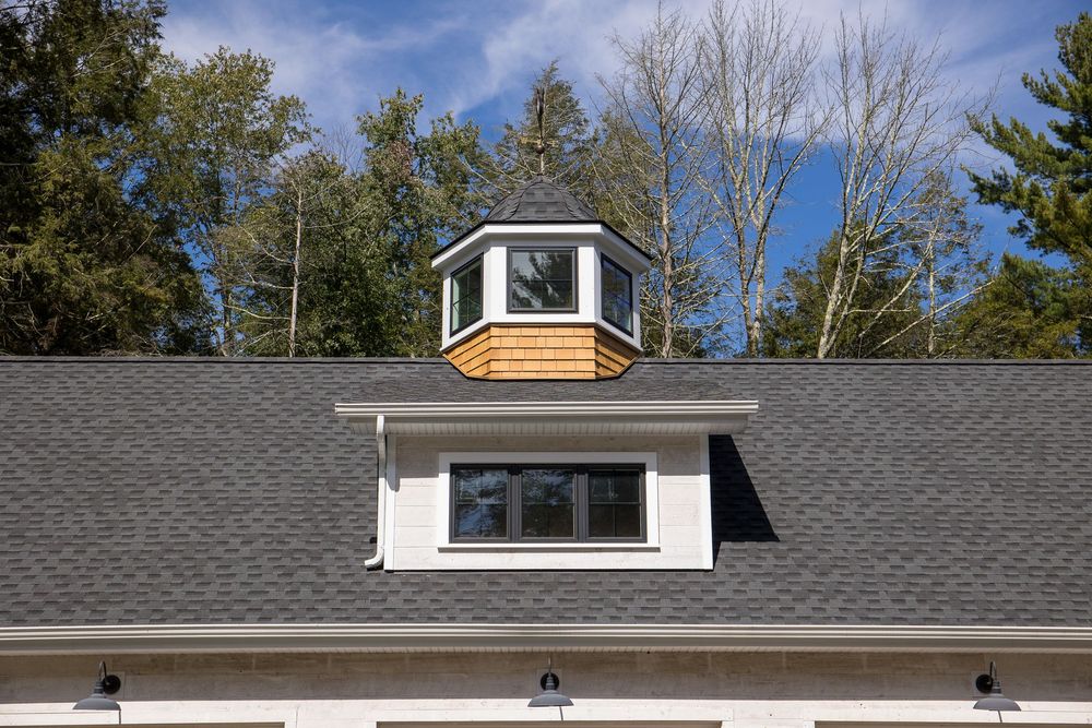 Decorative cupola with cedar shingles atop a white garage dormer with a large window and gray asphalt roof.