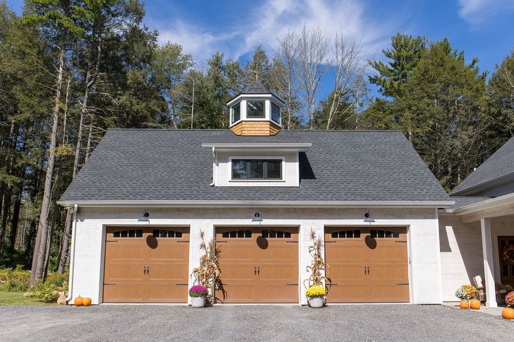 Three-car garage with wood-toned doors, white siding, cedar cupola, and fall harvest decorations under a blue sky.