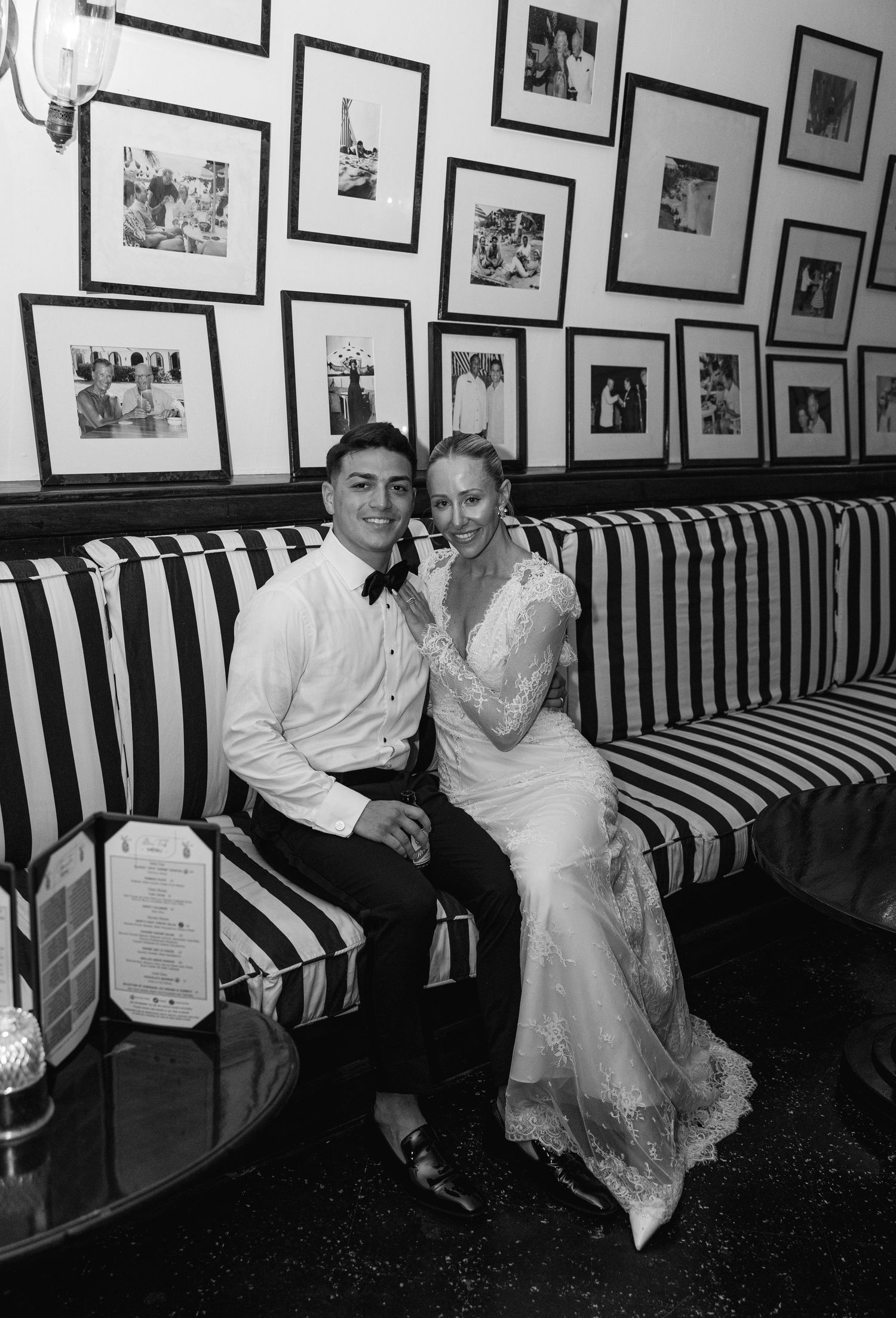 Black and white photo of a bride and groom sitting on a striped sofa, smiling, surrounded by framed photos on the wall.