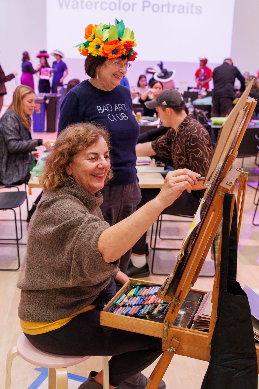 Woman painting at easel during watercolor portrait class, with instructor wearing flower crown observing her work.