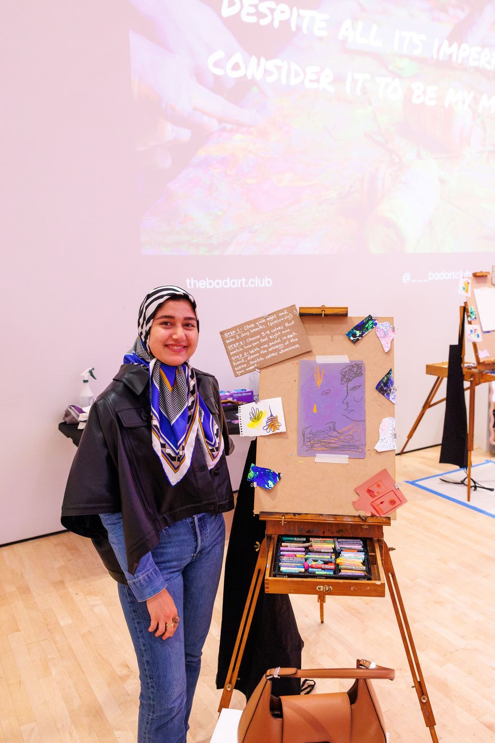 Young woman in hijab stands beside an art easel displaying colorful artwork at an indoor gallery exhibition.