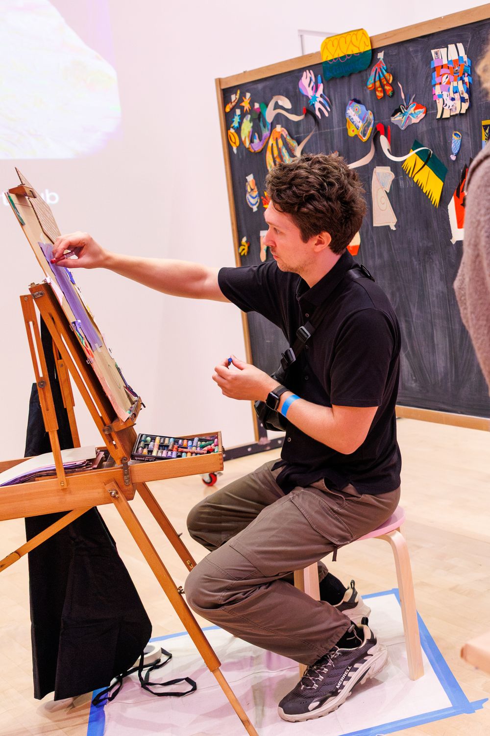 Artist painting on easel in studio with colorful chalk drawings on blackboard in background.