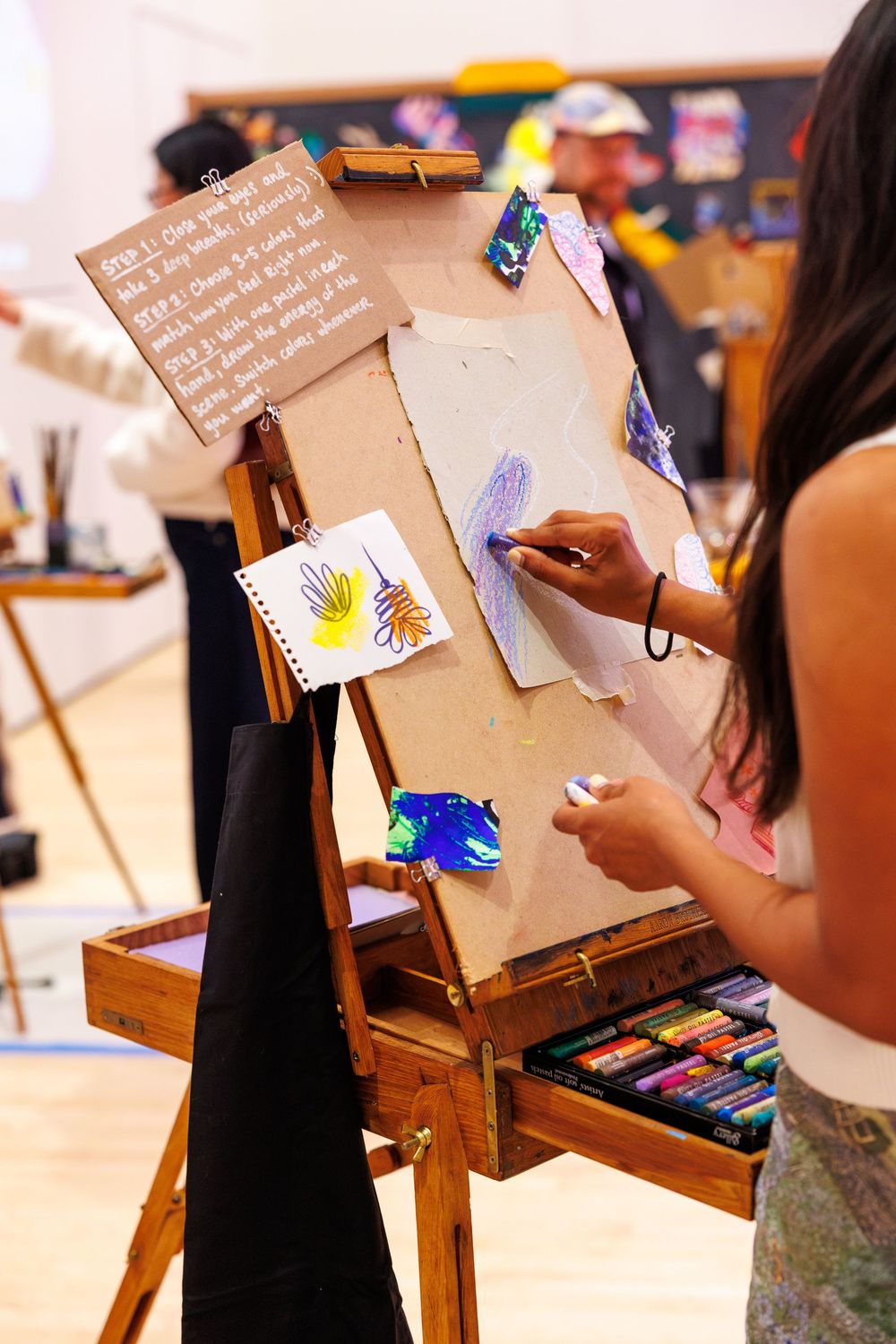 A student draws on canvas at a wooden easel with pastel crayons in an art class setting.
